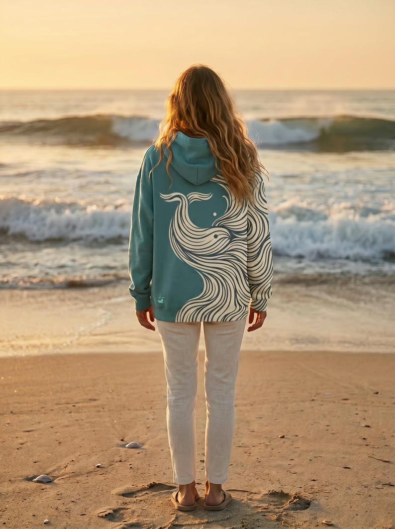 May include: A teal hoodie with a whale design on the back. The whale is depicted in white, flowing lines. The person is standing on a sandy beach, facing the ocean at sunset. The hoodie has a hood and long sleeves.