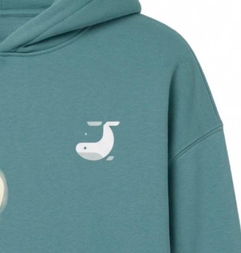 May include: Teal hoodie with a white whale graphic on the front. The hoodie has a hood and a relaxed fit. The whale graphic is a simple, minimalist design. The fabric appears to be a soft, comfortable material.
