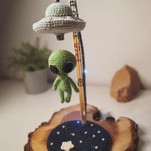 May include: A whimsical crochet lamp featuring a green alien figure suspended beneath a gray and green UFO. The lamp includes a dark blue base with white star accents, set on a wooden slice. The lamp is a unique decorative item.