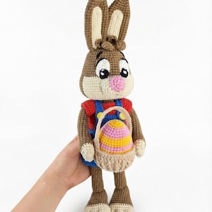 May include: A crocheted Easter bunny doll with brown and cream yarn. The bunny wears a red shirt and blue overalls, holding a basket with a colorful egg. The doll has large eyes, a pink nose, and long ears.