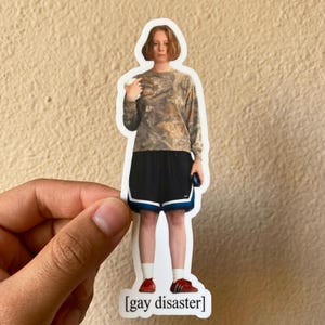 May include: A sticker featuring a person in a camouflage long-sleeve shirt, black shorts with a white and blue trim, and red shoes. The text '[gay disaster]' is at the bottom of the sticker.