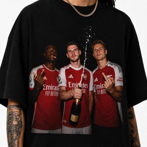 以下が含まれることがあります： 黒いTシャツ。赤と白のユニフォームを着た3人のサッカー選手がシャンパンボトルで祝うグラフィック。選手たちは中指を立てています。シャツには「Emirates Fly Better」の文字が入っています。