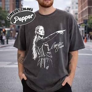 Puede incluir: Una camiseta gris carbón de manga corta con una ilustración gráfica blanca de una jugadora de baloncesto señalando hacia adelante, con el texto 'Comfort Colors Pepper' en la esquina superior izquierda y una firma junto a la jugadora.