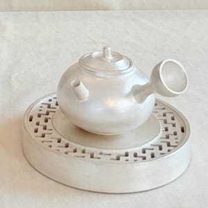May include: A white ceramic teapot with a distinctive spout and lid, set on a matching round base with a decorative pattern. The teapot has a minimalist, modern design, ideal for serving tea. The base measures approximately 15 cm in diameter.
