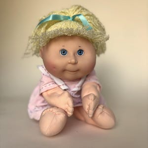 May include: A soft-sculpture doll with pale skin, blue eyes, and dimpled cheeks. The doll has short, curly yellow yarn hair tied with a light blue ribbon and wears a light pink dress with a white collar. The doll is seated with its feet forward.