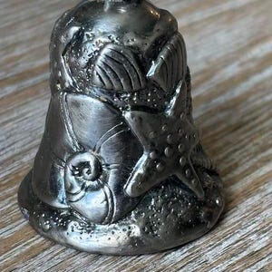 May include: A silver-toned metal bell ornament featuring a textured surface with raised relief designs of a starfish, a spiral seashell, and a fan-shaped scallop shell. The bell has a small loop at the top for hanging and a dark, weathered finish.