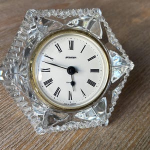 May include: A hexagonal clear crystal clock with a white face, black Roman numerals, and black hands. The clock features the brand name STANGER and the text QUARTZ and MADE IN GERMANY on the dial, surrounded by a gold-toned metal bezel.