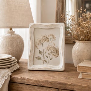 May include: A rectangular picture frame with a distressed white finish and a wavy, scalloped edge. The frame contains a botanical print featuring a delicate illustration of white hydrangea flowers with green leaves on a light cream background.