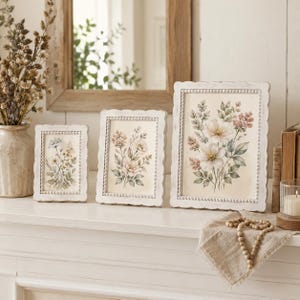 Puede incluir: Tres marcos de fotos rectangulares blancos con bordes de cuentas, cada uno con una impresión botánica de flores en tonos pastel suaves. Los marcos están dispuestos en fila sobre una superficie blanca, mostrando ilustraciones florales.