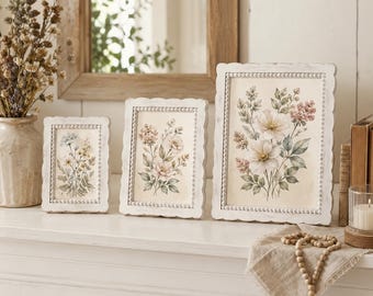 May include: Three rectangular white picture frames with beaded edges, each containing a botanical print of flowers in soft pastel tones. The frames are arranged in a row on a white surface, displaying floral illustrations with green leaves.