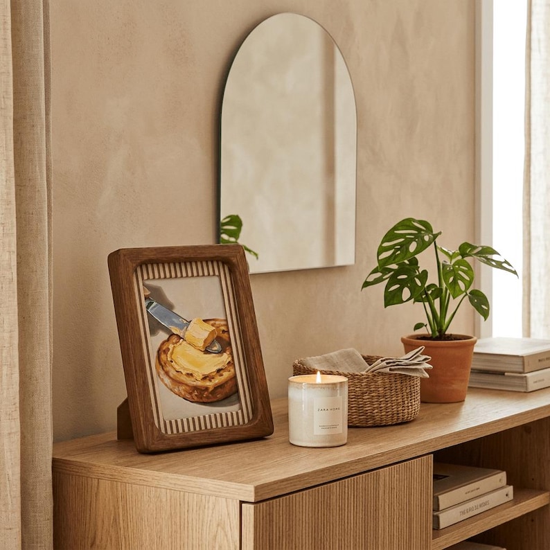 May include: A wooden framed picture of a knife spreading butter on a pastry, a lit candle, a small potted plant, and a woven basket on a wooden cabinet. An arched mirror hangs on the wall above the cabinet.