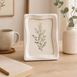 May include: A rectangular picture frame with a distressed white finish and a wavy, scalloped edge design. The frame contains a print of a green botanical illustration featuring delicate leaves and stems on a cream-colored background.