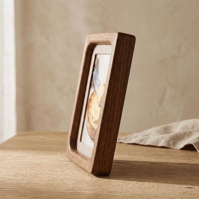 May include: A rectangular wooden picture frame with a dark brown finish. The frame has a thick border and a striped inner edge. The frame is angled, displaying a partial image of a painting. The frame is on a wooden surface.