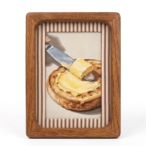 Puede incluir: Pintura enmarcada de una rebanada de pan tostado untada con mantequilla. Un cuchillo plateado extiende mantequilla amarilla sobre la tostada marrón. El marco es de madera marrón claro con borde a rayas. Tema de desayuno.