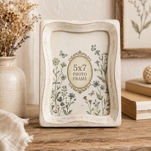 May include: A rectangular photo frame with a distressed white finish and curved edges. The insert features a central oval with the text 5x7 PHOTO FRAME, surrounded by a botanical illustration of green wildflowers and small butterflies on a cream background.