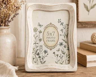 May include: A rectangular photo frame with a distressed white finish and curved edges. The insert features a central oval with the text 5x7 PHOTO FRAME, surrounded by a botanical illustration of green wildflowers and small butterflies on a cream background.