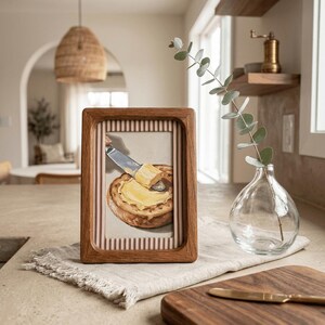 May include: A framed artwork depicts a slice of toasted bread being buttered with a knife. The frame is made of wood and has a striped background. The artwork is displayed on a neutral-colored surface with a small vase and cutting board.