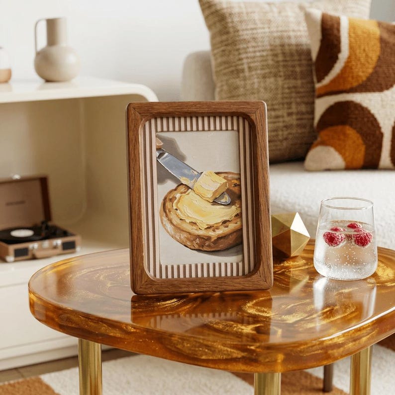 May include: A framed artwork depicts a slice of bread being buttered with a knife. The frame is made of wood with a striped border. The artwork is displayed on a glossy, gold-toned table with brass legs, alongside a glass of water with raspberries.