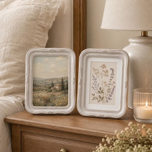 May include: Two rectangular picture frames with a distressed white finish and beaded detailing. The left frame displays a landscape painting of a countryside villa, while the right frame holds a botanical illustration of various wildflowers.