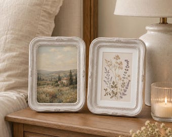 May include: Two rectangular picture frames with a distressed white finish and beaded detailing. The left frame displays a landscape painting of a countryside villa, while the right frame holds a botanical illustration of various wildflowers.