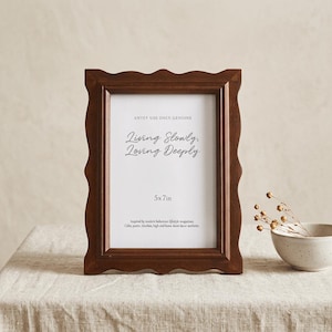 Puede incluir: Un marco de fotos de madera marrón oscuro con un borde ondulado, que contiene una impresión blanca con el texto "Living Slowly, Loving Deeply" y "5x7in". El marco está sobre un mantel de color neutro.