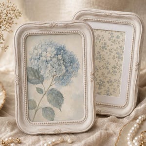 Op de afbeelding: Twee rechthoekige fotolijsten met een verweerde gebroken witte afwerking en gedetailleerde kralenranden. De ene lijst toont een aquarelillustratie van een blauwe hortensia, terwijl de andere een print met een klein blauw bloemenpatroon bevat.