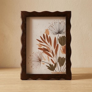Könnte beinhalten: Ein dunkelbrauner Bilderrahmen mit gewelltem Rand. Der Rahmen enthält einen Druck von botanischen Illustrationen in Braun-, Orange- und Grüntönen. Das Kunstwerk zeigt stilisierte Blumen und Blätter vor cremefarbenem Hintergrund. Der Rahmen steht auf einer Holzoberfläche.