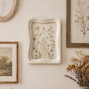 May include: A rectangular wall frame with a distressed white finish featuring a wavy, contoured edge. Inside the frame is a botanical print displaying various wildflowers, small leaves, and stems in muted yellow, green, and soft purple tones.