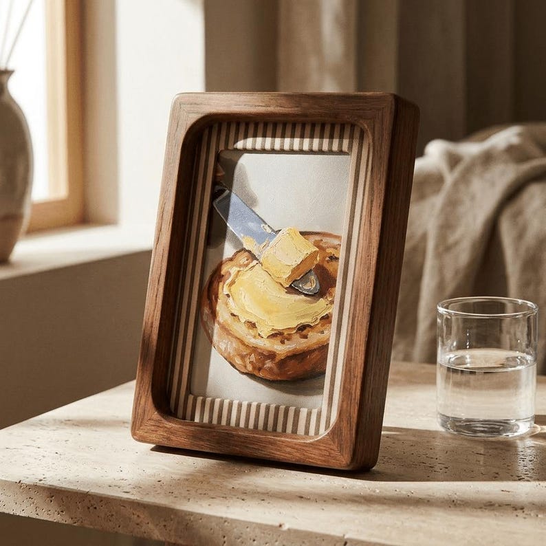 May include: A wooden picture frame with a painting of a piece of toast with butter being spread on it. The frame is a warm brown color and has a striped background. A glass of water sits to the right of the frame.