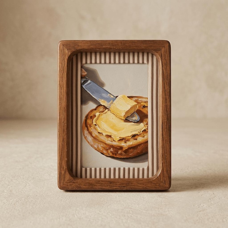 May include: A framed artwork depicts a slice of toasted bread being buttered with a knife. The painting features a wooden frame with a light-colored background and vertical stripes. The butter is a bright yellow color.