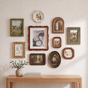 May include: A gallery wall arrangement featuring various picture frames in wood, gold, and green finishes, displaying black and white family photos, botanical prints, and landscape art, positioned above a light wood console table with a ceramic vase.