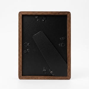 May include: A rectangular wooden picture frame with a dark brown finish. The back of the frame is black, with a black easel stand and metal clips to hold the backing in place. The frame is designed to hold a photograph or artwork.