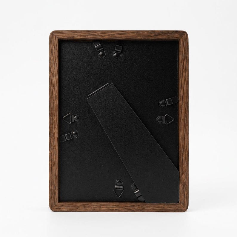 May include: A rectangular wooden picture frame with a dark brown finish. The back of the frame is black, with a black easel stand and metal clips to hold the backing in place. The frame is designed to hold a photograph or artwork.