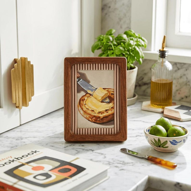 May include: A framed painting depicts a knife spreading butter on a toasted English muffin. The frame is brown with a striped border. A bowl of limes, a cookbook, and a bottle of olive oil are also visible.