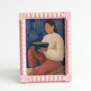 Könnte beinhalten: Gerahmtes Gemälde mit einem rosa Perlenrahmen. Das Kunstwerk zeigt eine Person, die eine blaue Schüssel hält und ein weißes Hemd und rostfarbene Hosen trägt. Der Hintergrund ist dunkelblau.