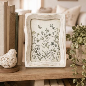 May include: A rectangular picture frame with a distressed white finish featuring a botanical print of green wildflowers and small butterflies on a cream background. The frame has a wavy, contoured edge and stands upright on a wooden surface.