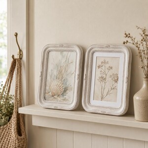 Op de afbeelding: Twee rechthoekige fotolijsten met een verweerde gebroken witte afwerking en kralenranden aan de binnenzijde. De linkerlijst bevat een aquarel van een schelp en helmgras, terwijl de rechterlijst een botanische print van een bloeiende plant toont.