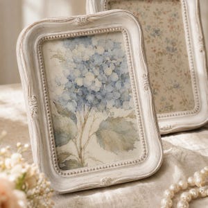 Op de afbeelding: Een rechthoekige fotolijst met een verweerde gebroken witte afwerking en een kralenrand aan de binnenzijde. De lijst bevat een aquarelprint van blauwe hortensia's met groene bladeren op een cr&egrave;mekleurige achtergrond, geplaatst in een grotere lijst.