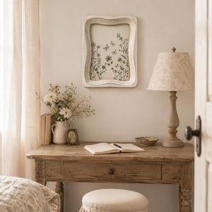 May include: A rectangular white wooden wall frame with a wavy edge, featuring a botanical illustration of wildflowers and butterflies in black ink on a cream background, mounted on a wall above a wooden desk with a lamp and a small stool.