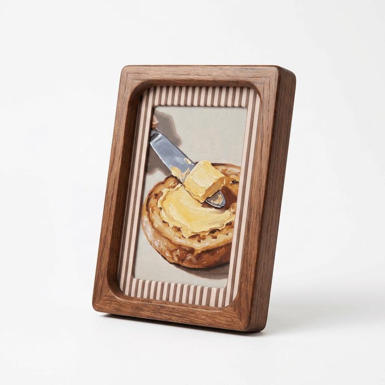 May include: A framed painting depicts a slice of bread with butter being spread on it. The painting is in a wooden frame with a striped border. The butter is yellow, and the bread is golden brown.