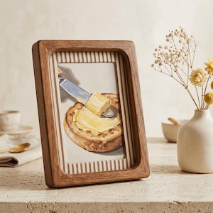May include: A framed artwork depicts a slice of toasted bread with butter being spread on it. The artwork is in a wooden frame with a striped border. A small white vase with dried flowers sits to the right of the frame.