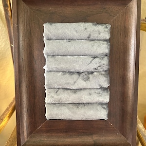May include: A rectangular dark brown wooden frame containing six horizontal rows of soft, light gray velvet fabric. The velvet is arranged in padded, cylindrical strips stacked vertically within the center of the wooden frame structure.