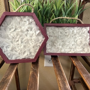 Puede incluir: Dos adornos de pared con marcos de madera color granate oscuro e inserciones de piel sintética blanca. Un marco es hexagonal y el otro rectangular. Cada pieza tiene un lazo de cuerda de yute para colgar, mostrando un patrón texturizado y arremolinado en la tela de felpa blanca.