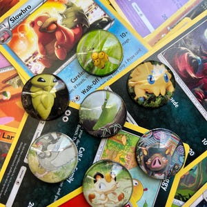 May include: A collection of six circular glass cabochons featuring various colorful creature illustrations, arranged on top of a scattered pile of trading cards with visible text including Slowbro, Will-O-Wisp, and Careless.