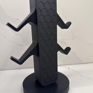 K&ouml;nnte beinhalten: Schwarzer Gaming-Headset-St&auml;nder mit rundem Sockel und vertikaler St&uuml;tze mit Wabenmuster. Der St&auml;nder hat vier abgewinkelte Arme zum Halten von Headsets. Das Design ist modern und elegant.