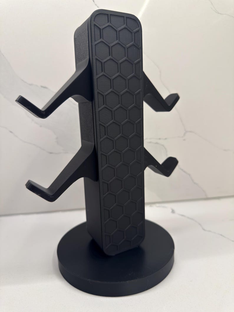K&ouml;nnte beinhalten: Schwarzer Gaming-Headset-St&auml;nder mit rundem Sockel und vertikaler St&uuml;tze mit Wabenmuster. Der St&auml;nder hat vier abgewinkelte Arme zum Halten von Headsets. Das Design ist modern und elegant.