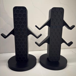 K&ouml;nnte beinhalten: Zwei schwarze Hantelst&auml;nder. Jeder St&auml;nder hat einen rechteckigen Mitteltr&auml;ger mit Wabenmuster und drei Paaren abgewinkelter Arme zum Halten von Hanteln. Die St&auml;nder stehen auf runden Sockeln.