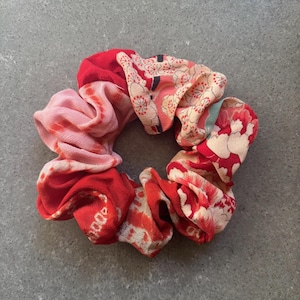 Könnte beinhalten: Ein farbenfrohes Haargummi mit einer Mischung aus roten, rosa- und weißen Blumenmustern. Der Stoff ist gerafft und gefaltet, wodurch eine strukturierte, kreisförmige Form entsteht. Dieses Haaraccessoire setzt Farbakzente.