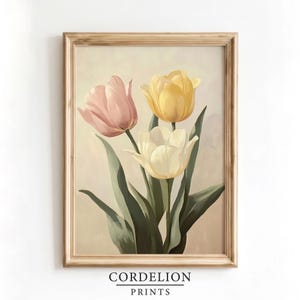 Op de afbeelding: Een ingelijste kunstdruk met drie tulpen in zachte pasteltinten van roze, geel en wit, tegen een neutrale beige achtergrond. De bloemen hebben lange groene stelen en bladeren, alles gevat in een lichtbruine houten lijst.