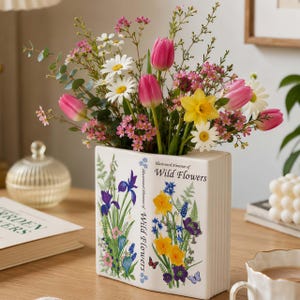 Puede incluir: Un jarrón de cerámica blanco con forma de libro que presenta ilustraciones botánicas de lirios morados, narcisos amarillos y flores azules con el texto "Illustrated Almanac of Wild Flowers". El jarrón contiene un ramo de tulipanes rosas, margaritas blancas, narcisos amarillos y eucalipto verde.